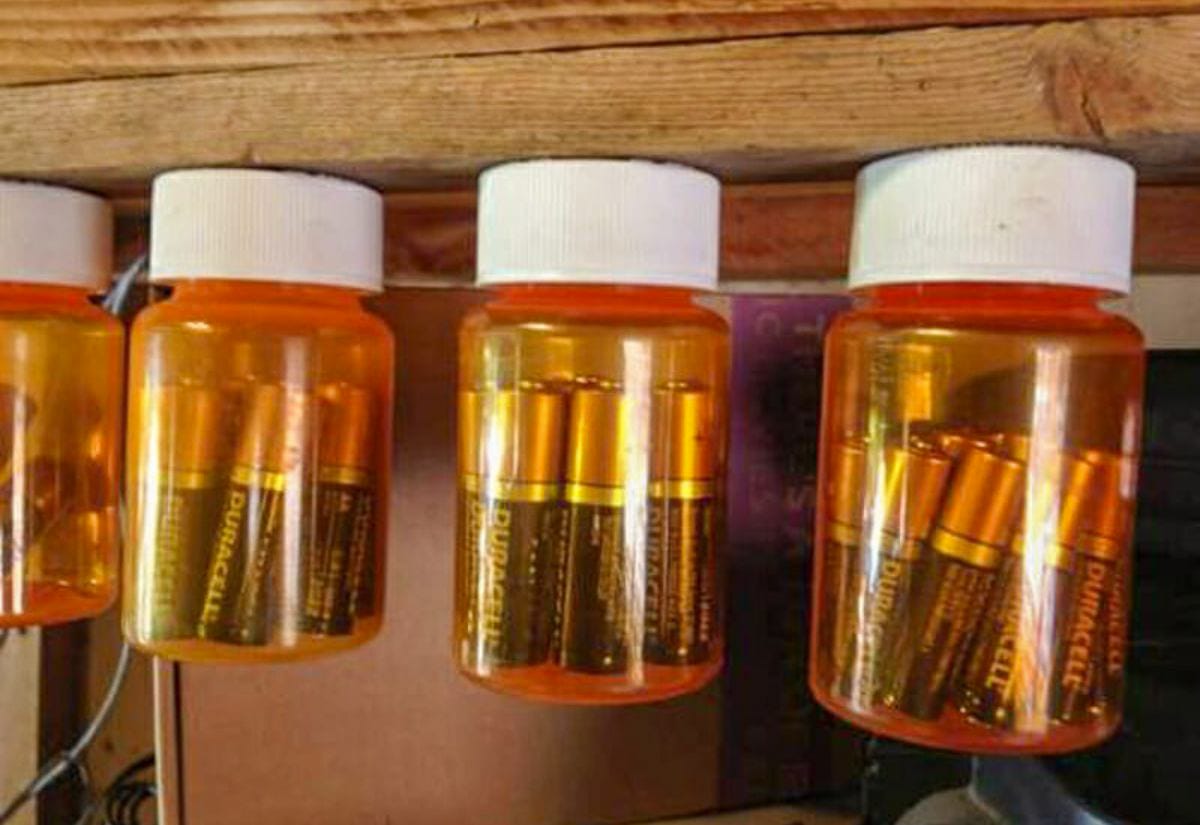 Pill bottles mounted under a wooden shelf, each filled with AA batteries for organized storage.