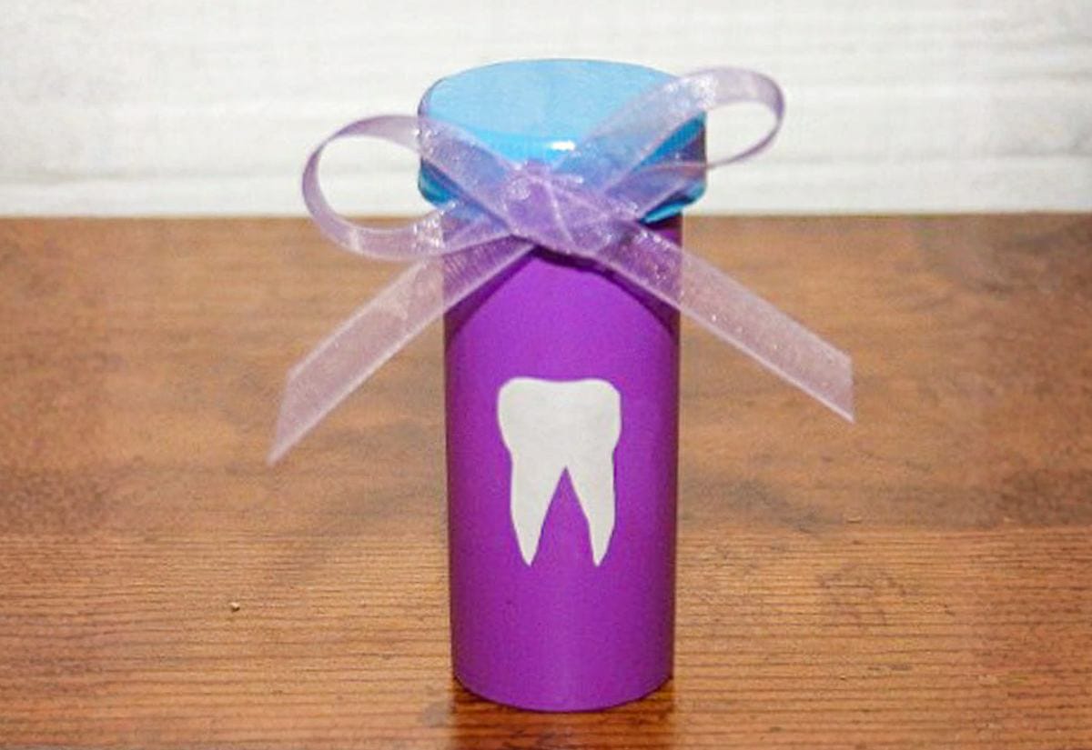 Purple pill bottle with a white painted tooth design, blue cap, and pink ribbon bow, used as a tooth storage container.