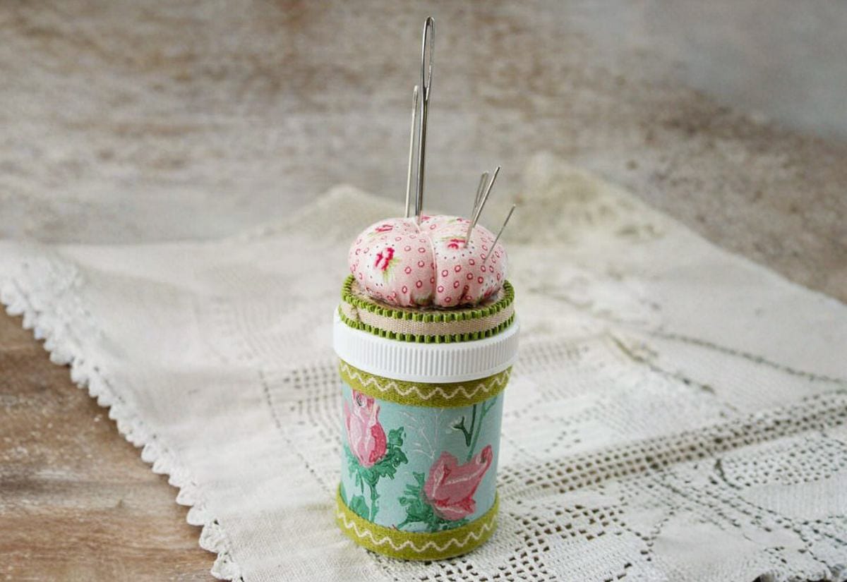 Prescription pill bottle decorated with fabric and trim, topped with a pincushion and sewing needles to make a portable sewing kit.