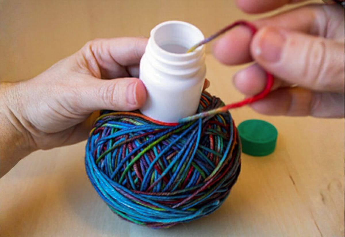 Hands pulling yarn through a hole in a pill bottle cap while winding a center-pull yarn ball.