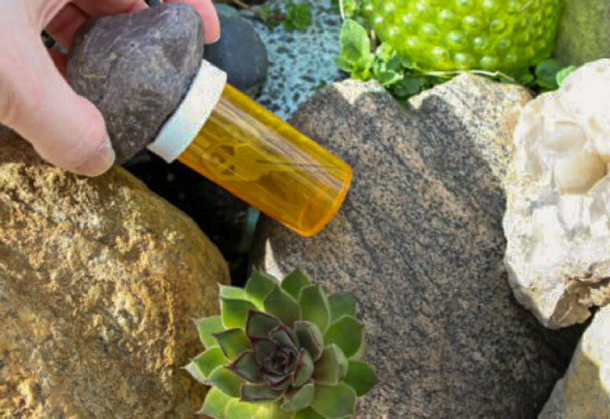 Hand holding a pill bottle with a rock glued to the lid, tucked among outdoor landscaping stones as a hidden key holder.