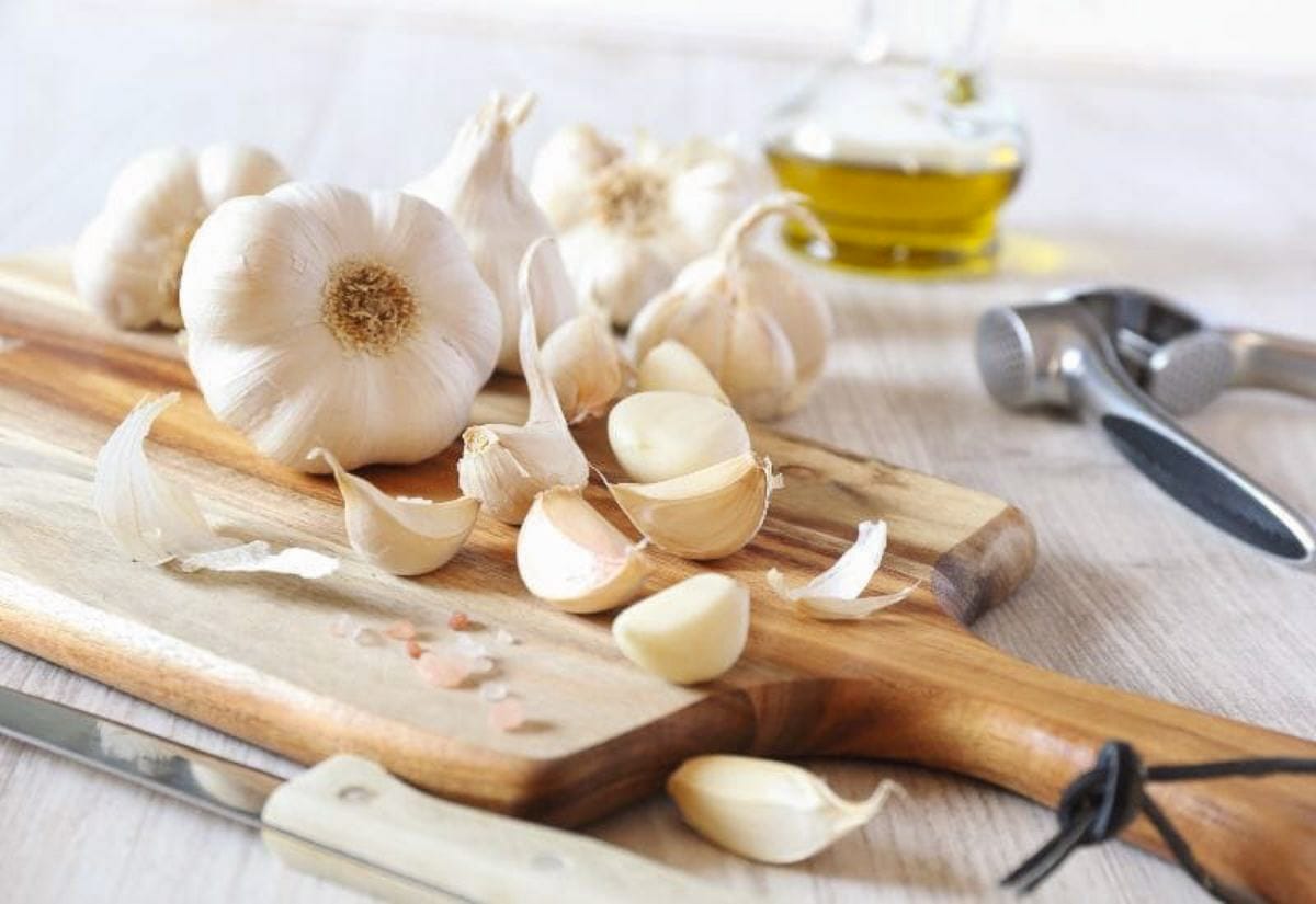 Peeled garlic cloves on a wooden cutting board with garlic press and olive oil in the background, step one in how to preserve garlic for cooking.