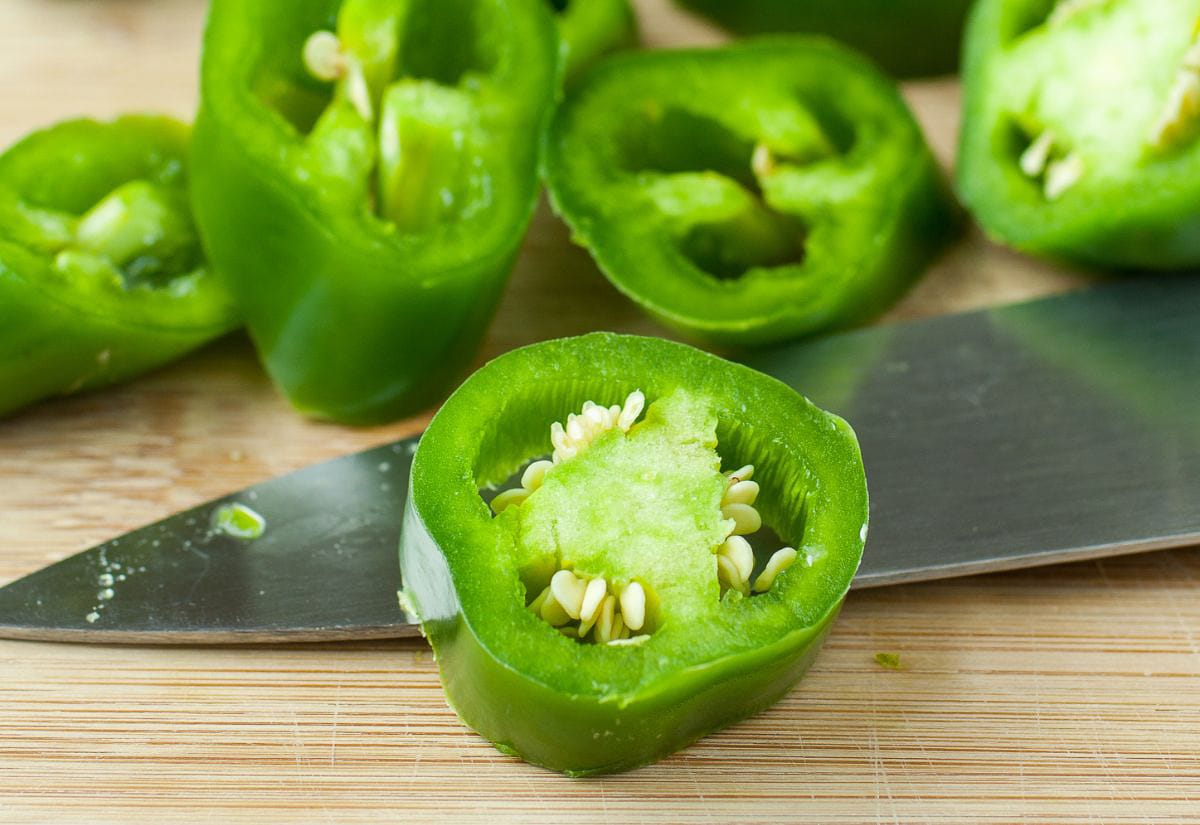 Fresh jalapeños sliced into rings on a cutting board beside a knife, step one in how to preserve hot peppers.