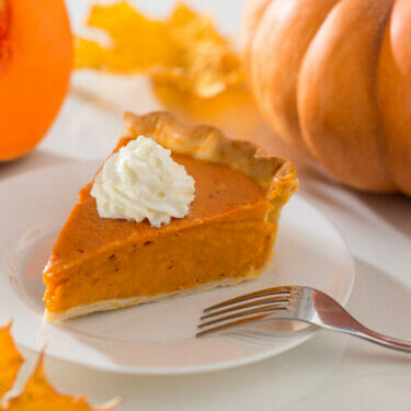A slice of pumpkin pie with a dollop of whipped cream and pumpkins in the background.