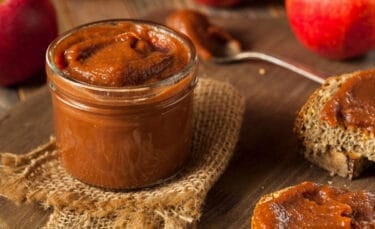 A glass jar of thick, dark brown crabapple butter on a piece of burlap, surrounded by apples and toast spread with butter.