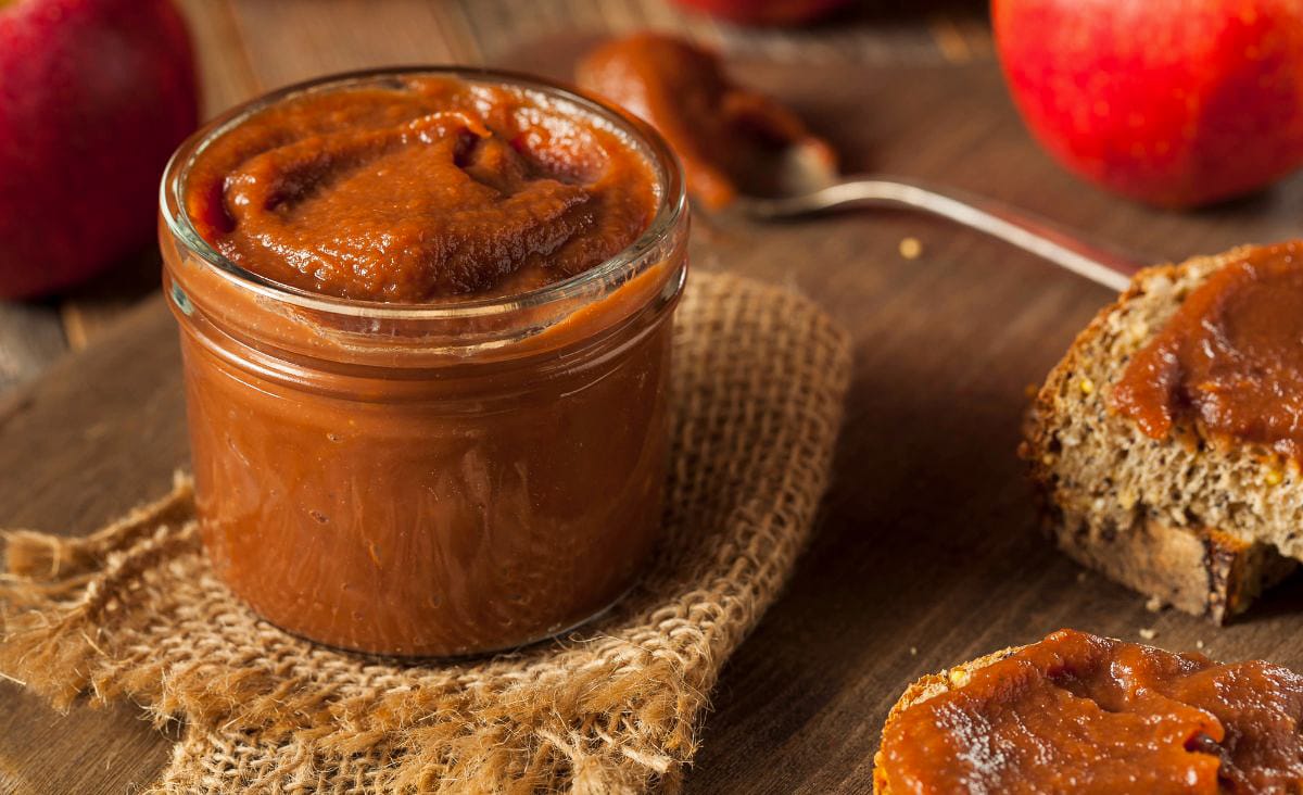 A glass jar of thick, dark brown crabapple butter on a piece of burlap, surrounded by apples and toast spread with butter.