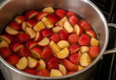 Quartered red crabapples with skins simmering in a stainless steel pot of water on a kitchen stove.