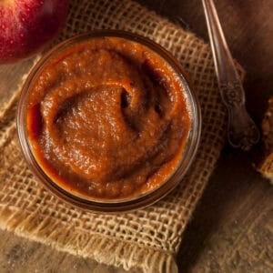 A rustic jar of smooth, homemade crabapple butter on a wooden surface beside an apple and vintage spoon.