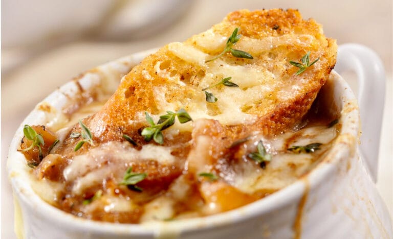 Close-up of French onion soup in a white bowl, showing the caramelized onions, melted cheese, and toasted bread with fresh thyme leaves on top.