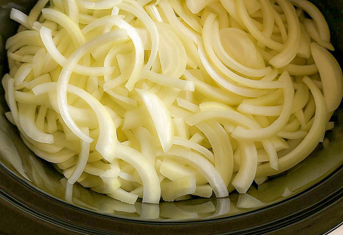 Thinly sliced yellow onions piled inside a black slow cooker before cooking, lit by soft, natural kitchen light.