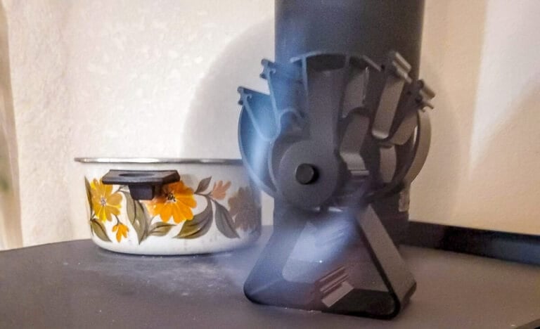 Close-up of a heat-powered stove fan spinning beside a vintage enamel pot on a wood stove, demonstrating how the fan runs using only the stove’s heat.
