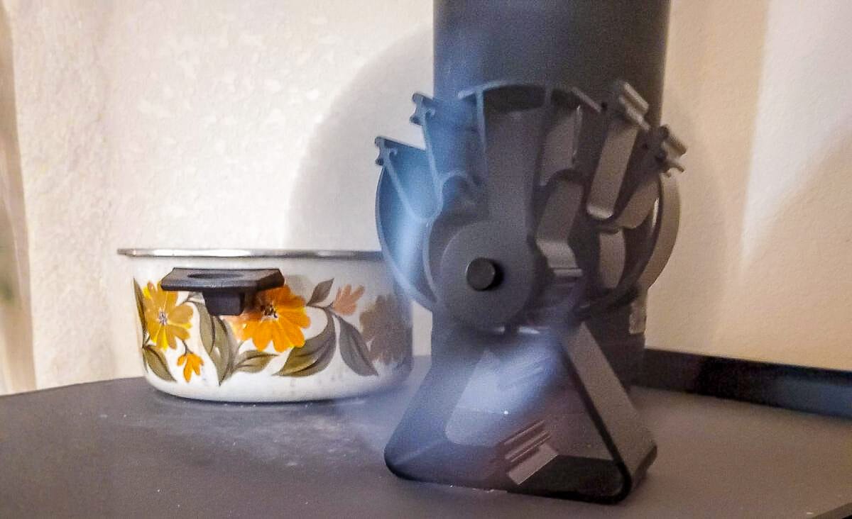 Close-up of a heat-powered stove fan spinning beside a vintage enamel pot on a wood stove, demonstrating how the fan runs using only the stove’s heat.