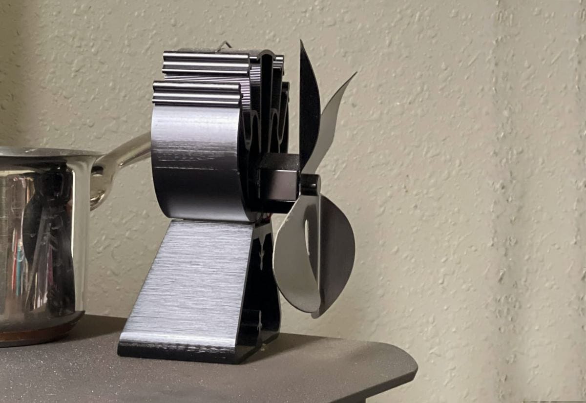 Side view of a modern heat-powered stove fan running beside a metal saucepan on a wood stove, displaying the fan’s design and heat-activated operation.
