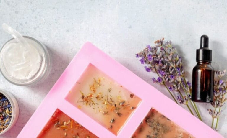 Close-up of a pink silicone soap mold filled with melt-and-pour soap and dried botanicals, surrounded by lavender sprigs and small jars on a light countertop.