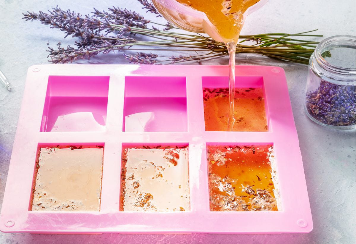 Melted soap being poured from a measuring cup into a pink silicone mold, with dried lavender stems and a jar of lavender buds arranged in the background.
