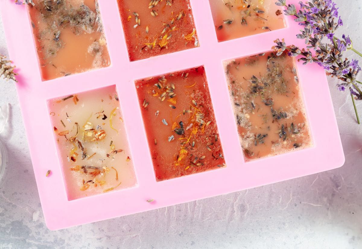 A pink silicone mold holding several melt-and-pour soap bars infused with dried flowers and herbs, with lavender sprigs placed beside the mold on the countertop.