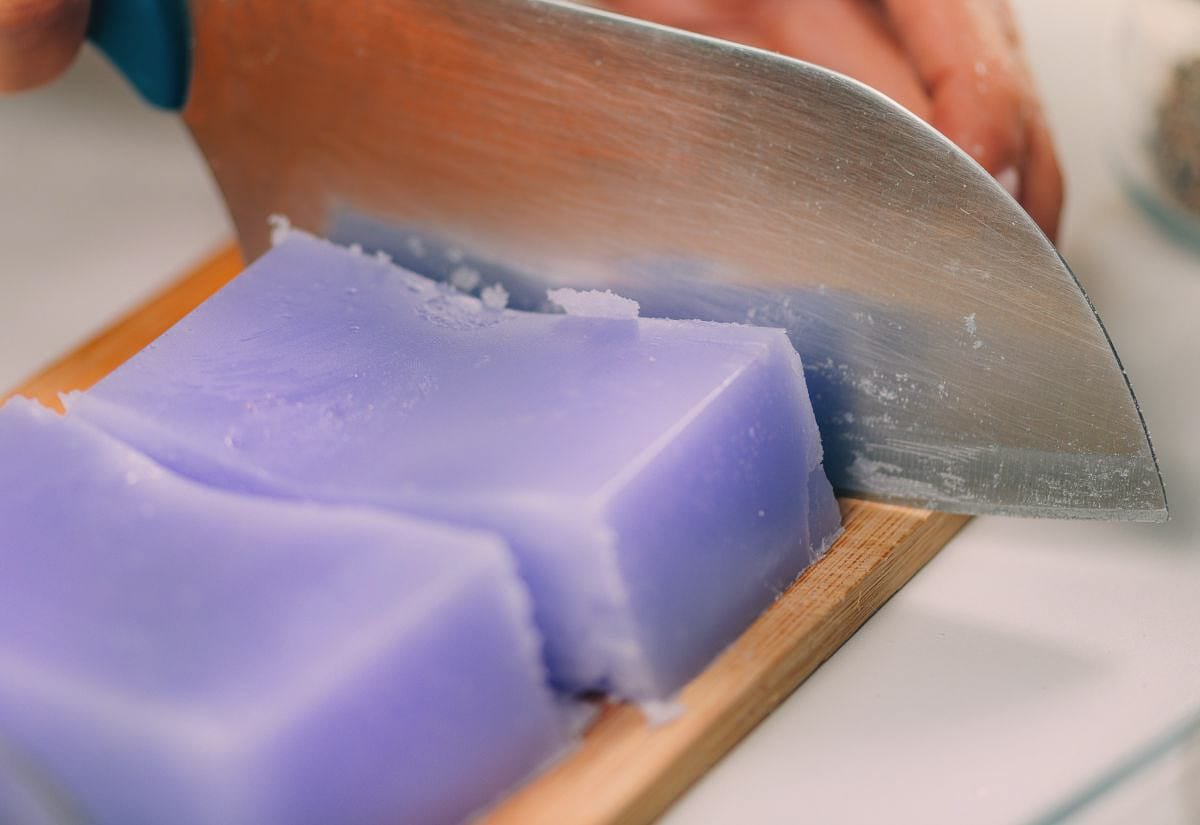 A block of purple melt-and-pour soap being sliced on a wooden cutting board with a large kitchen knife.