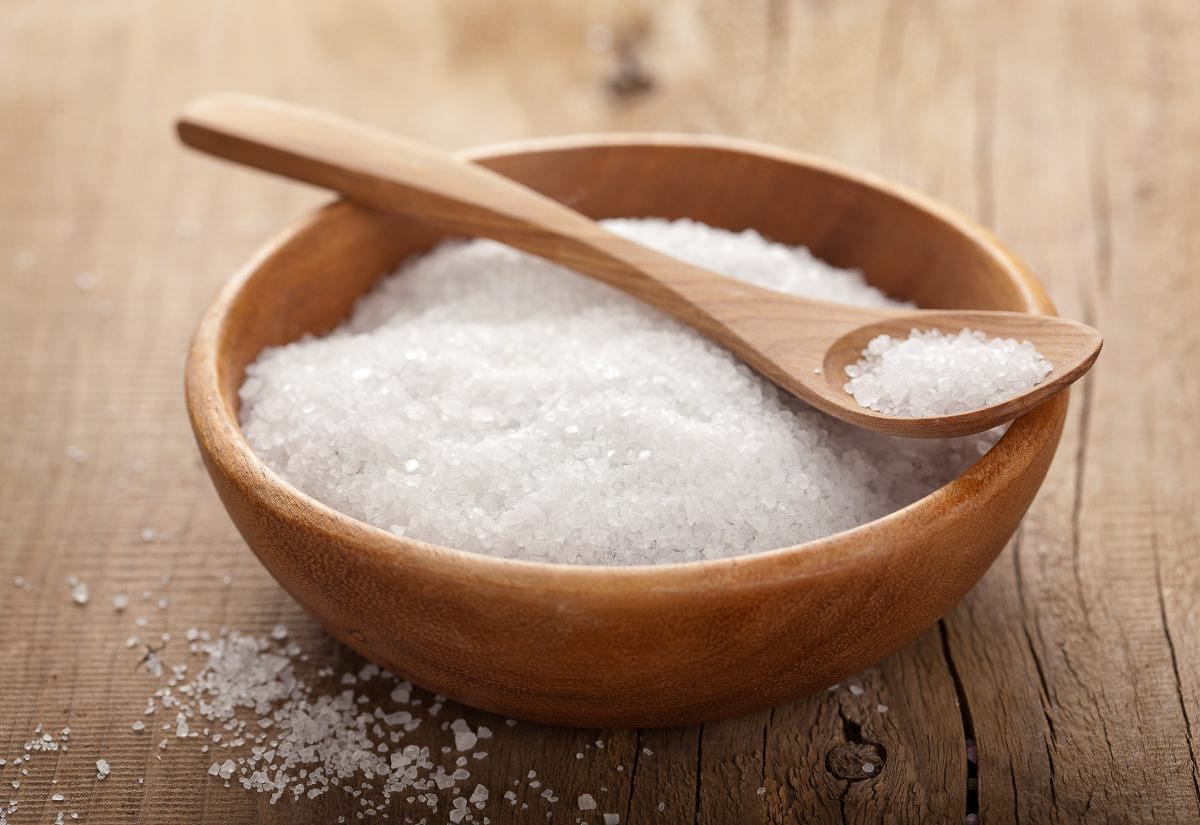Wooden bowl filled with coarse white Epsom salt on a rustic wooden surface, with a wooden spoon resting inside the bowl and loose salt crystals scattered around.