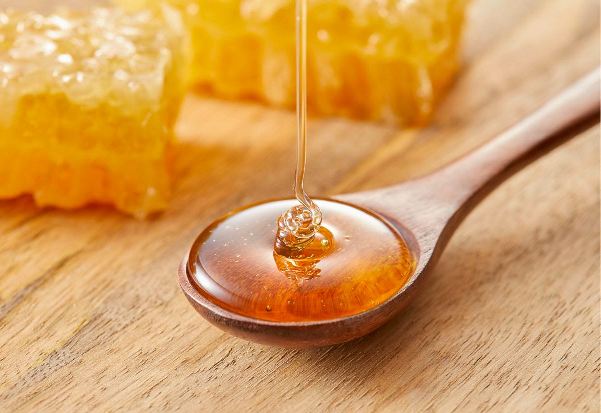 Close-up of golden honey drizzling from a dipper into a wooden spoon, with honeycomb pieces blurred in the background on a wooden surface.