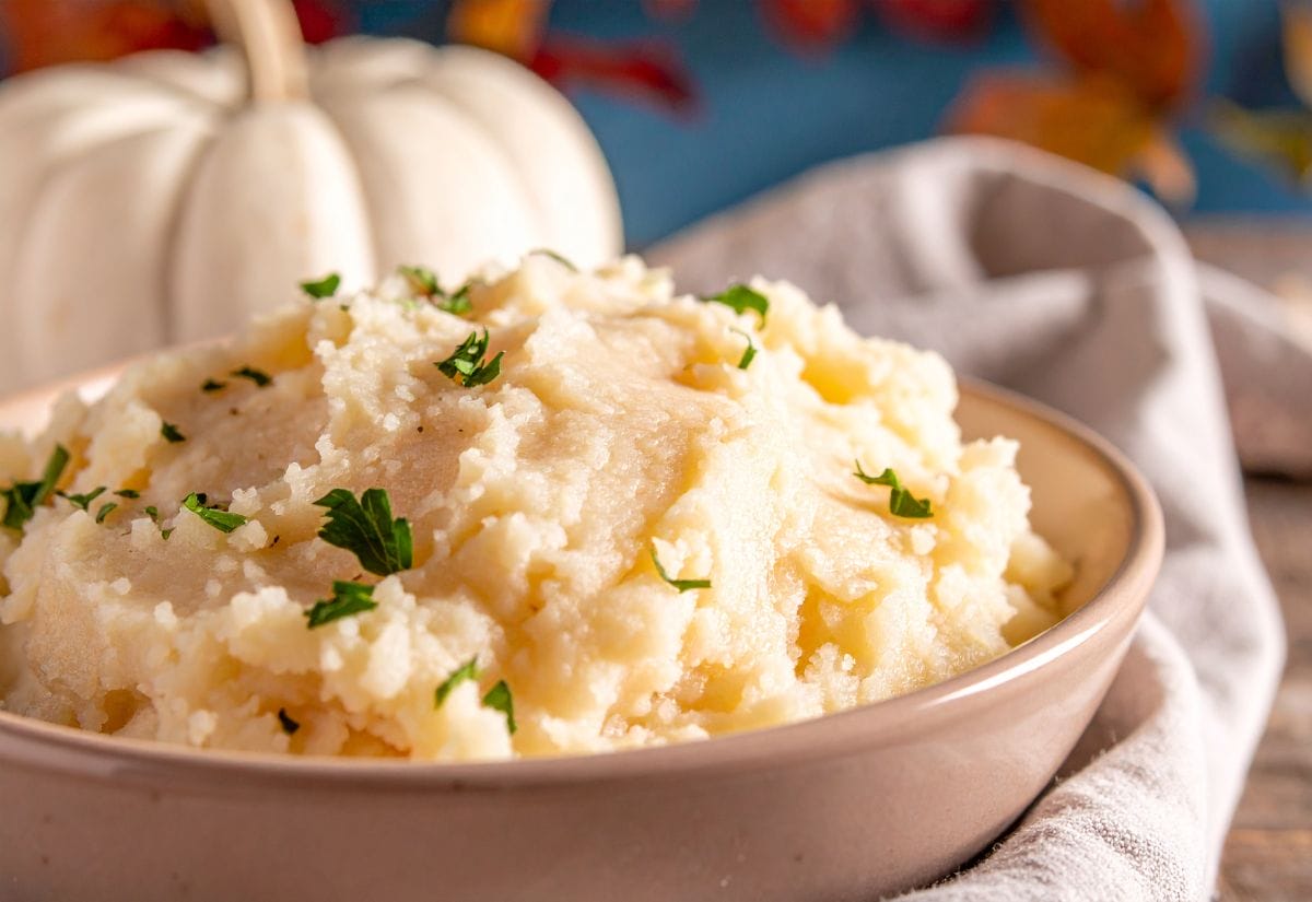 A warm bowl of homemade mashed potatoes topped with melted butter and fresh parsley, sitting on a rustic table beside a white pumpkin — cozy Thanksgiving side dish ready to serve.