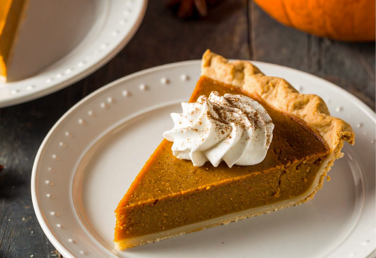 A slice of pumpkin pie with a flaky golden crust and a swirl of whipped cream dusted with cinnamon, served on a white plate — a classic Thanksgiving dessert.