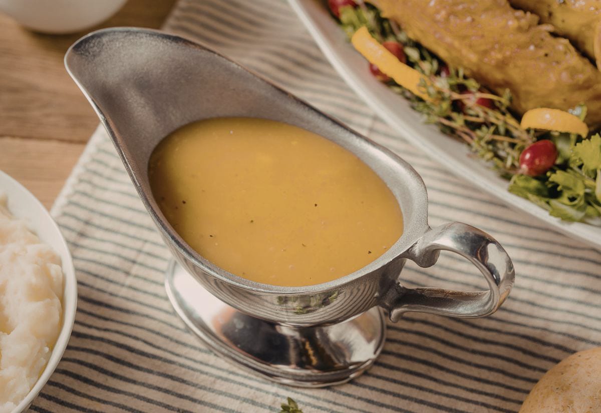 Silver gravy boat filled with rich homemade turkey gravy, resting on a striped cloth next to a bowl of mashed potatoes — part of a cozy Thanksgiving spread.