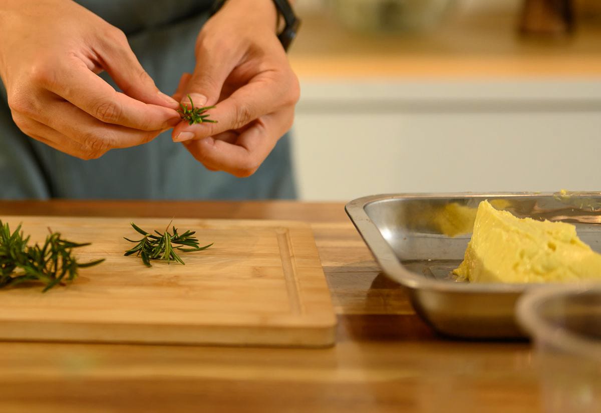 Hands preparing fresh rosemary sprigs on a wooden cutting board beside a pan of homemade compound butter — simple make-ahead prep for Thanksgiving cooking.