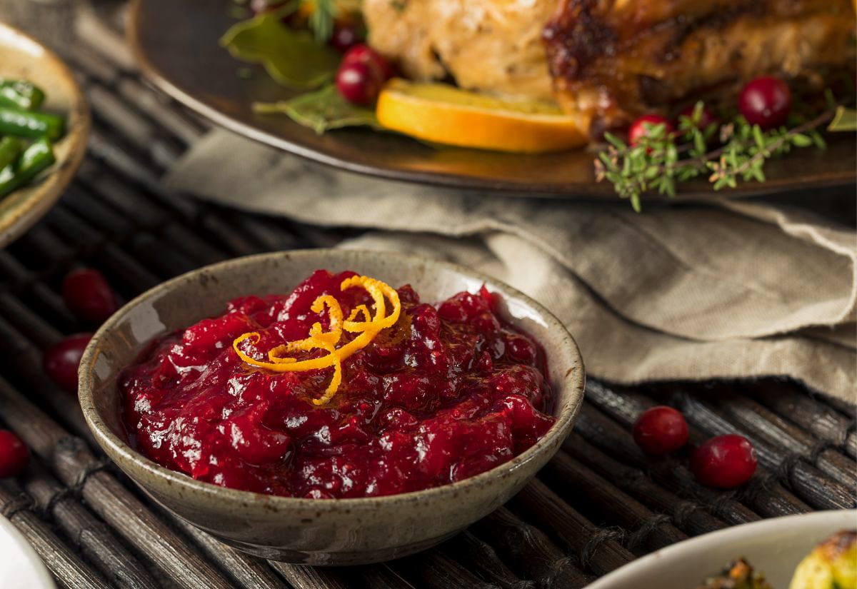 Homemade cranberry sauce with orange zest garnish in a rustic bowl, surrounded by Thanksgiving dishes and fresh cranberries — vibrant, tangy holiday favorite.