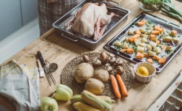 Uncooked turkey in a roasting pan beside chopped vegetables, potatoes, and herbs on a wooden counter — early Thanksgiving prep before cooking begins.