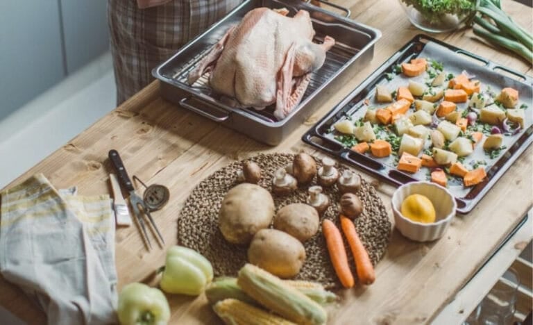 Uncooked turkey in a roasting pan beside chopped vegetables, potatoes, and herbs on a wooden counter — early Thanksgiving prep before cooking begins.