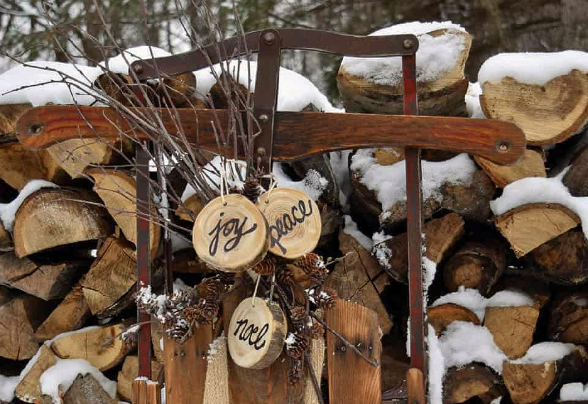 An old wooden sled propped in front of a snowy woodpile, decorated with twigs, pinecones, and wood-slice ornaments that read joy, peace, and noel.