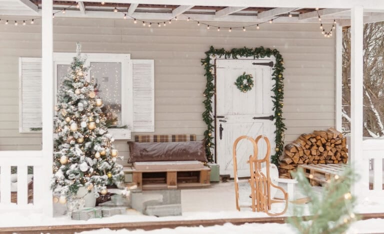 Cozy winter porch with a lit Christmas tree, garland-wrapped white door, string lights overhead, stacked firewood, and a wooden sled leaning near the entry.