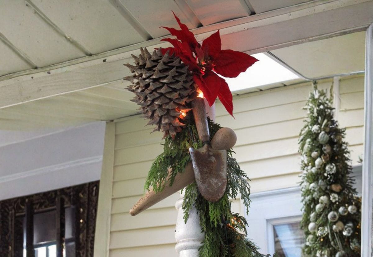 Rusty garden trowels and hand tools used as outdoor Christmas decor, bundled with evergreen branches, a large pinecone, and a red poinsettia on a porch column.