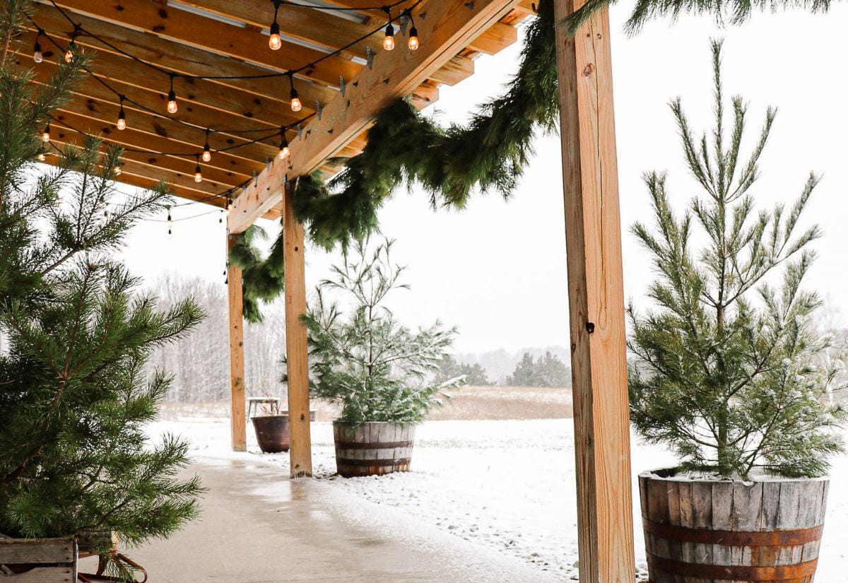 Fresh evergreen garland and warm string lights decorating a wooden porch overhang, with small Christmas trees in wooden barrels along the walkway.