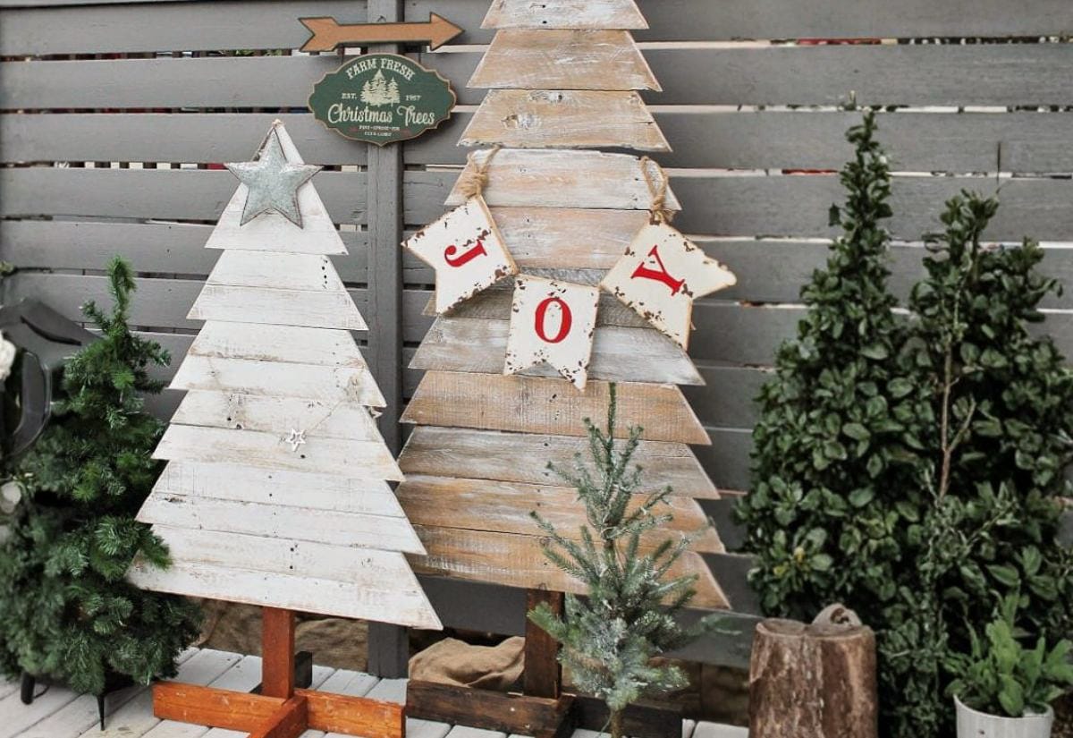Rustic outdoor display of handmade wooden pallet Christmas trees painted white and natural wood, surrounded by small evergreens and vintage-style holiday decor.