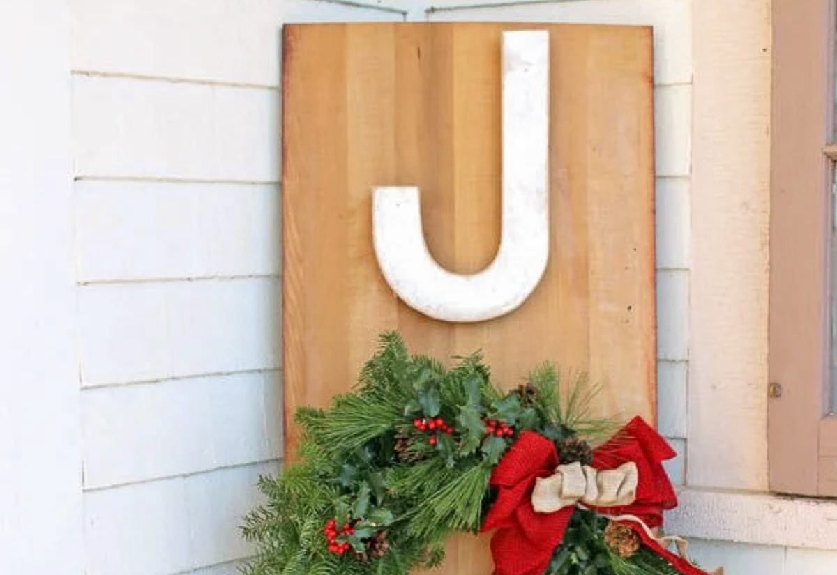 A tall wooden porch sign with a large white letter J and Y and a fresh evergreen wreath decorated with red berries, pinecones, and a red bow as the O in JOY.