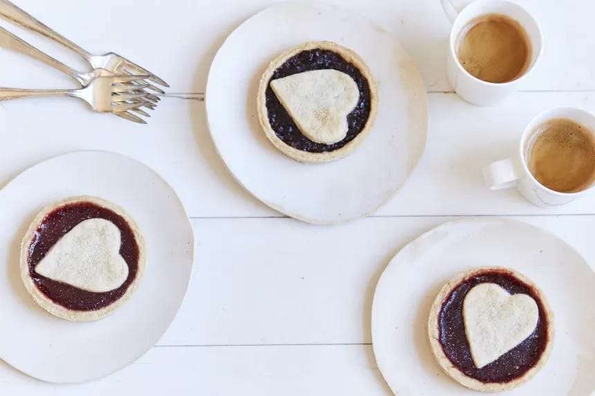 Jam tarts with a heart of crust on the top, on a white plate with cups of coffee and forks.