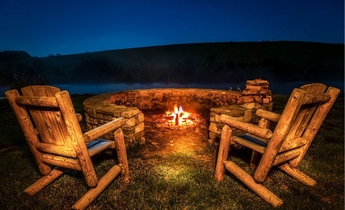 Two wooden adirondack chairs positioned in front of a campfire and stone wood ring.