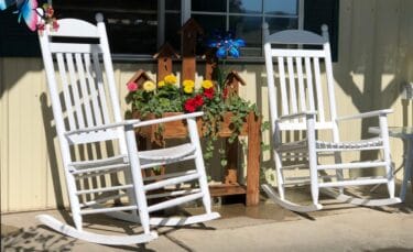 Two rocking chairs on a farmhouse porch with planters filled with flowers.