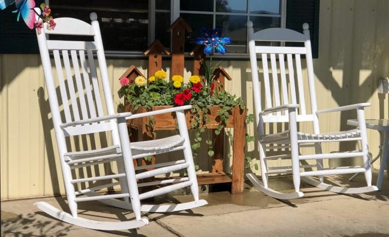 Two white rocking chairs on a sunny porch beside a rustic flower planter and birdhouse decor.
