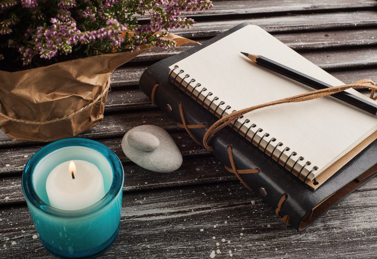A cozy setup with a lit candle, journal, pencil, and a potted plant on a weathered wooden table.