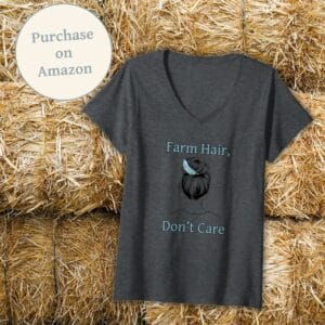 A dark gray Farm Hair, Don’t Care v-neck t-shirt displayed against a background of stacked hay bales. The shirt features a trendy messy bun with a feather accent, making it a stylish and comfortable choice for farm life enthusiasts. A "Purchase on Amazon" badge is included to guide customers to the product listing.