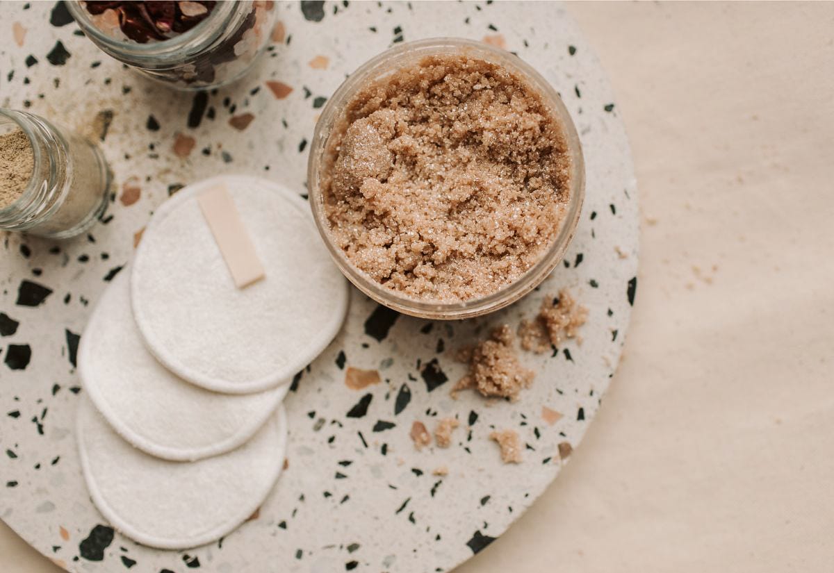 An open jar of homemade brown sugar scrub on a speckled tray, surrounded by reusable cotton rounds and small jars of natural ingredients.