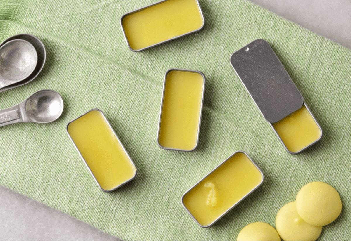 Several small metal tins filled with yellow homemade lip balm, arranged on a green cloth with measuring spoons nearby.