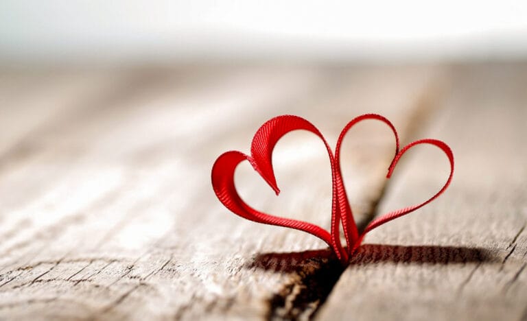 Red satin ribbon shaped into two small hearts resting on a weathered wooden surface, with soft natural light creating gentle shadows.