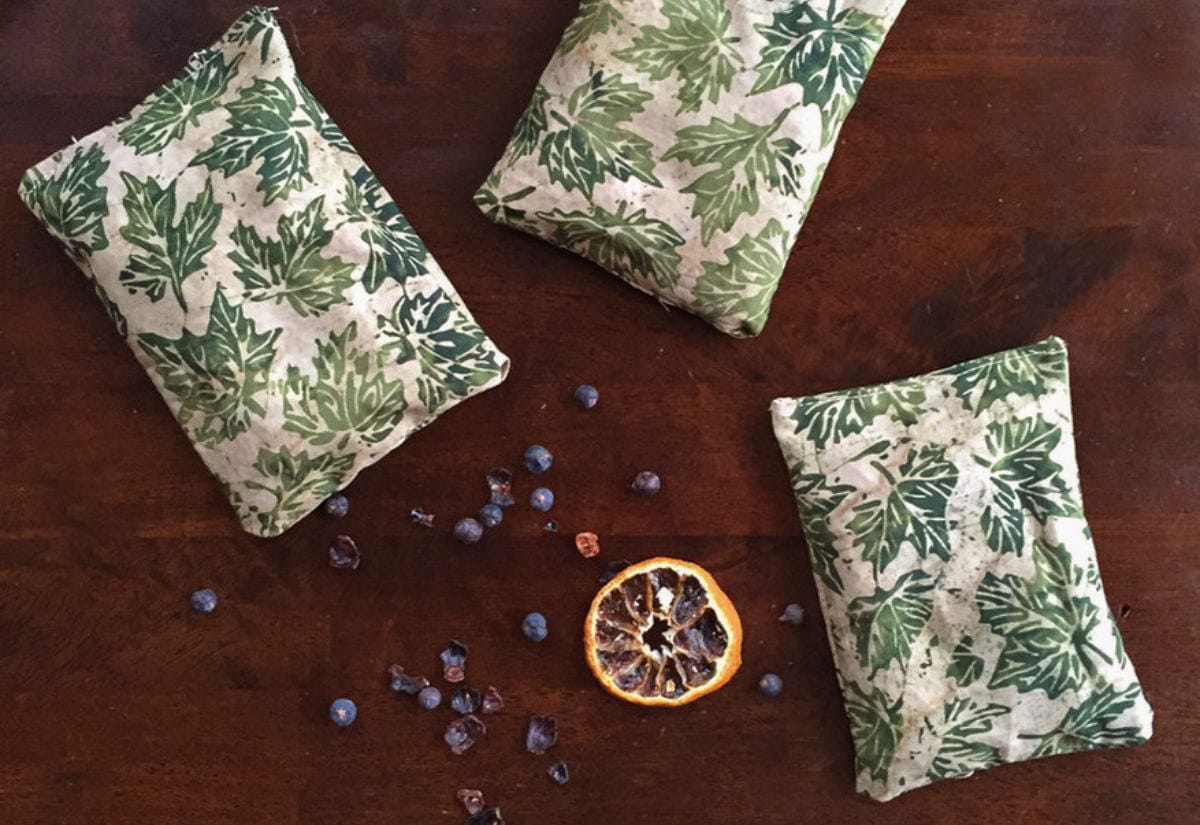 Three handmade fabric sachets with green leaf prints arranged on a dark wooden table, surrounded by dried juniper berries and a slice of dried orange.