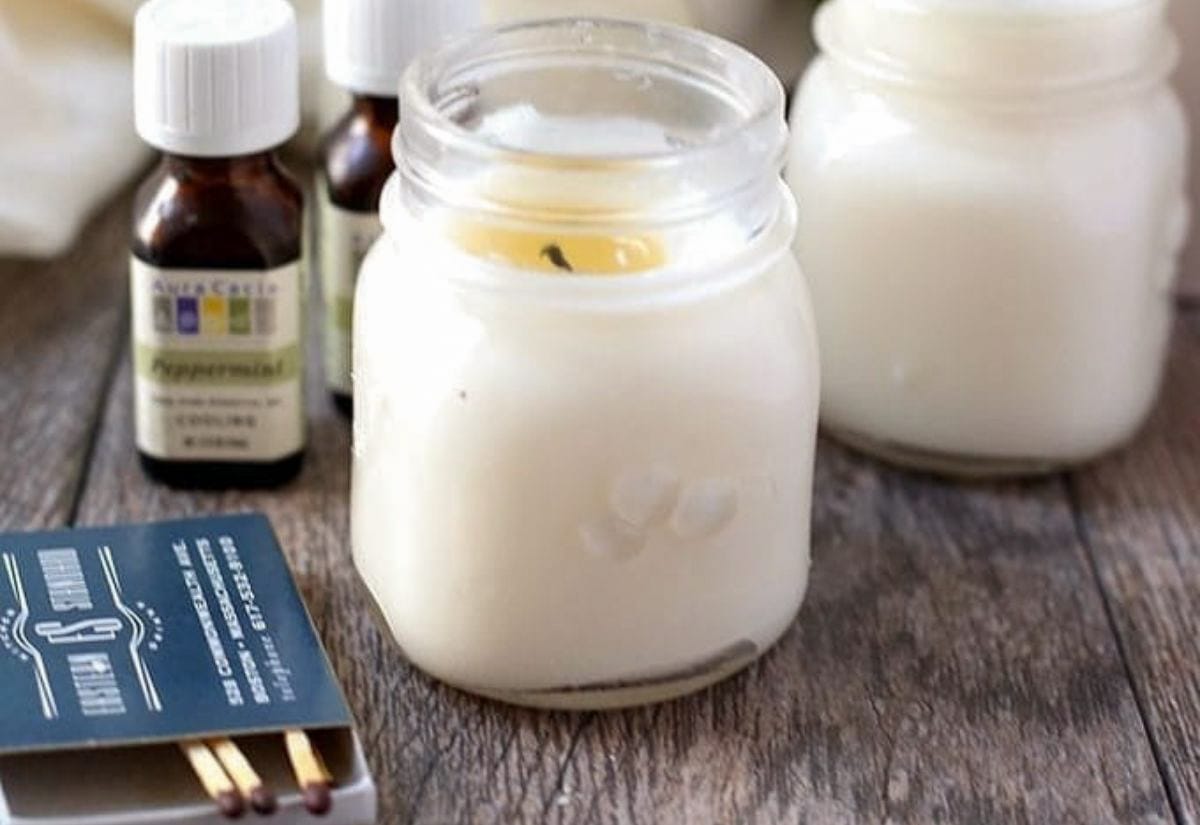 Small mason jar candles filled with creamy white wax, sitting on a rustic wood surface alongside essential oil bottles and an open matchbox.