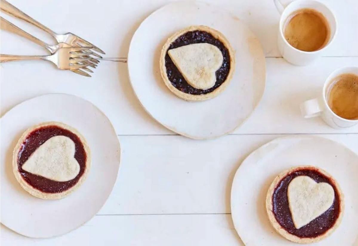 Three small jam tarts topped with heart-shaped pastry cutouts, plated on simple white dishes with cups of coffee and silver forks nearby.
