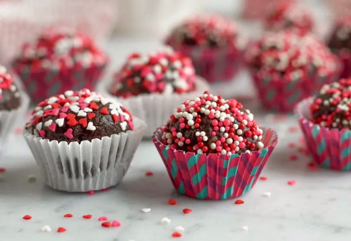 Chocolate truffles coated in red, white, and pink heart sprinkles, arranged in colorful paper cups on a light countertop.