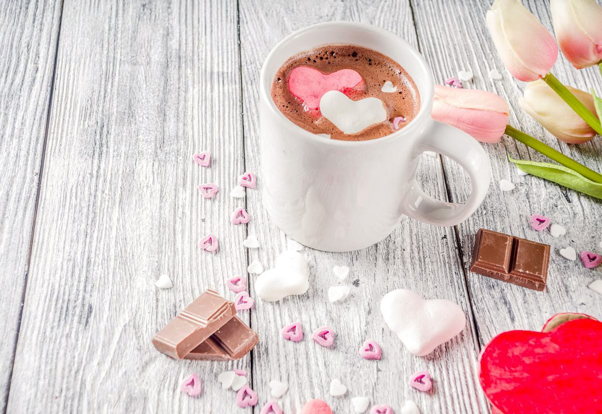 A white mug of hot chocolate topped with pink and white heart-shaped marshmallows, surrounded by chocolate pieces, heart sprinkles, and pink tulips on a gray wood surface.